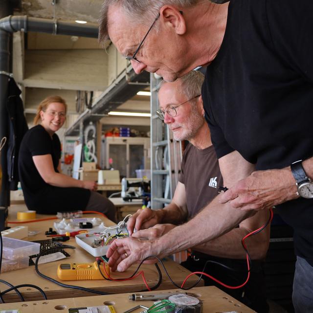 Eine Foto aus der Hanswerkstatt. Zu sehen sind zwei ältere Personen die ein Messgerät an einer defekten Lampe anlegen. Im Hintergrund schaut eine jüngere Person den beiden bei der Arbeit zu.